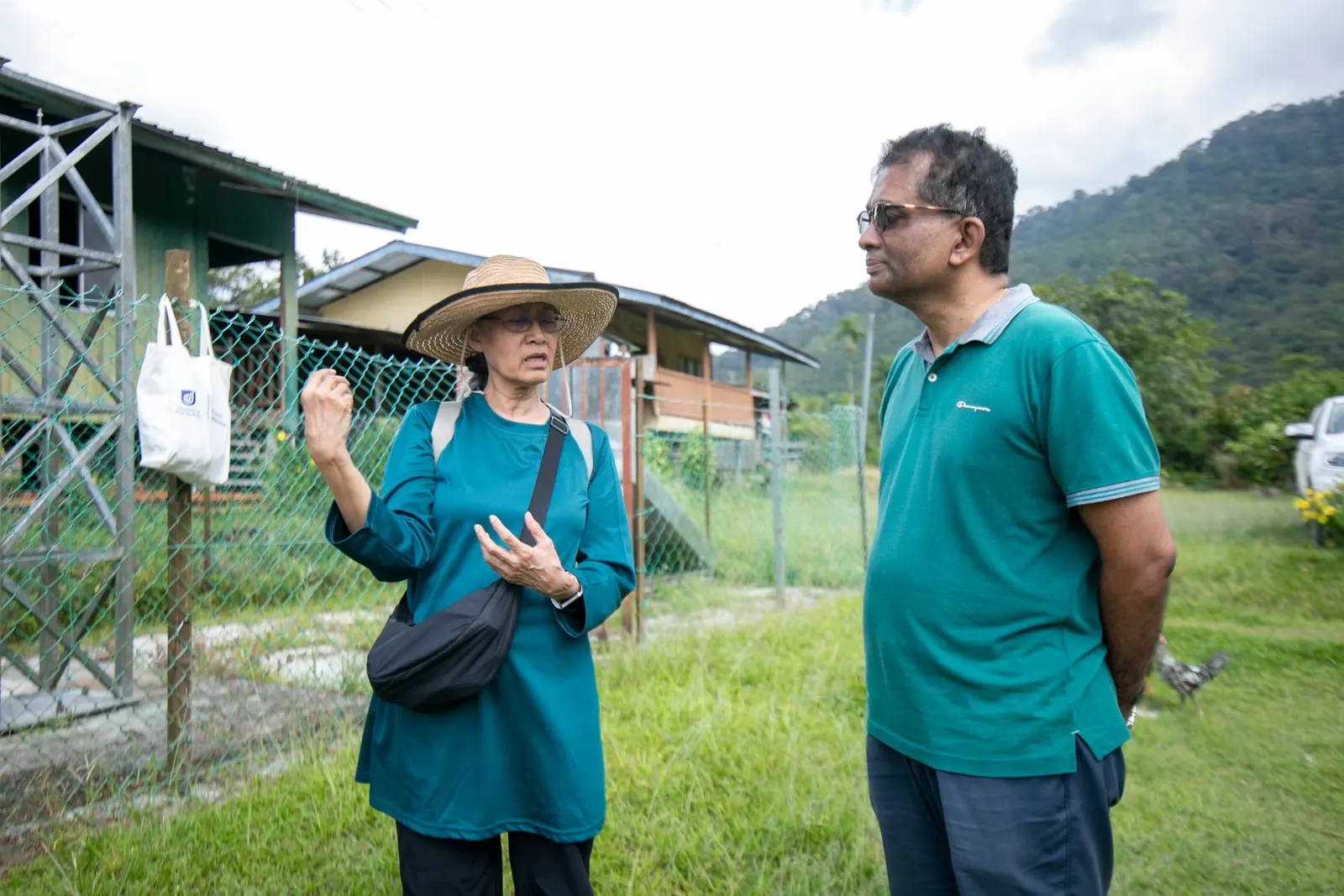 Alice Lasimbang, in a wide straw hat, talking with Nithi Nesadurai on the grass beside village buildings and forested hills