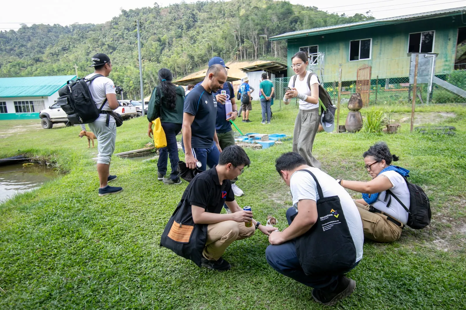 Members of the group gathered on the grass in Buayan Village, some crouching to greet a puppy, with village houses and jungle behind