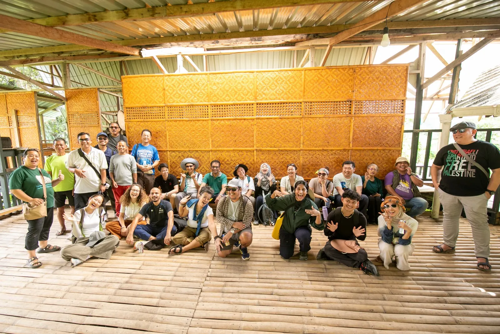 The full CANSEA group photographed together on the bamboo floor of an open-sided village hall with woven walls