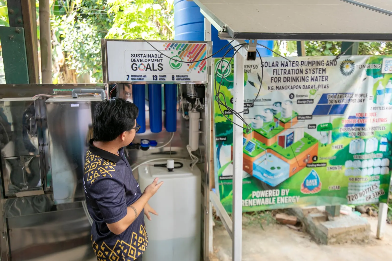 Adrian Lasimbang beside a solar-powered water filtration and dispensing unit, with an explanatory banner about the system behind it