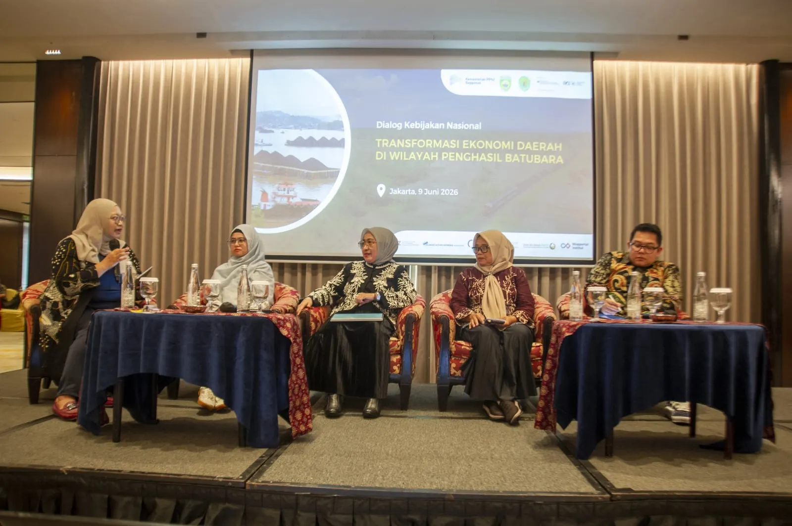Five panellists seated on stage beneath the dialogue's title slide, one speaking into a microphone
