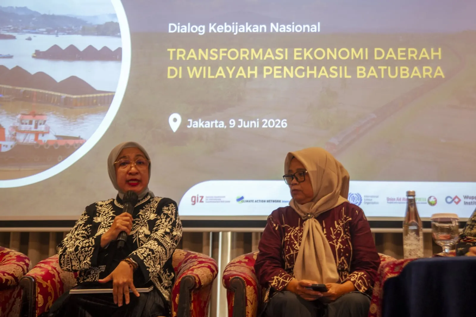 Two women panellists seated together, the one on the left speaking into a microphone, the title slide behind them