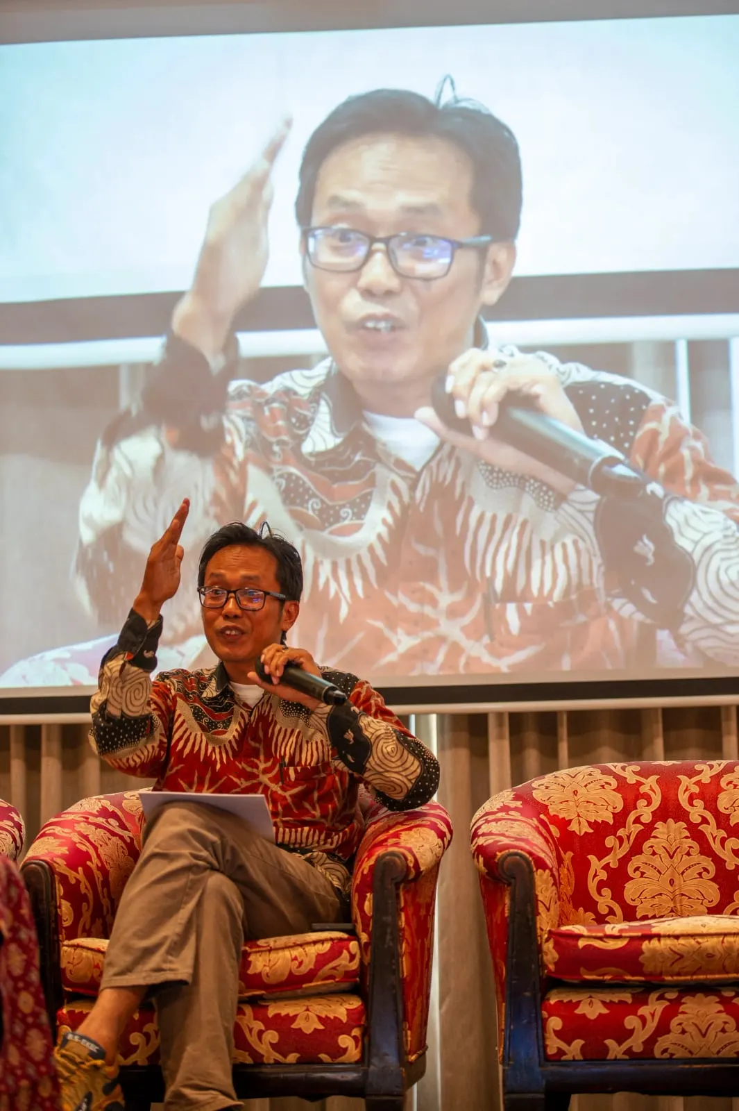 A panellist speaking into a microphone with one hand raised, his image projected large on the screen behind him