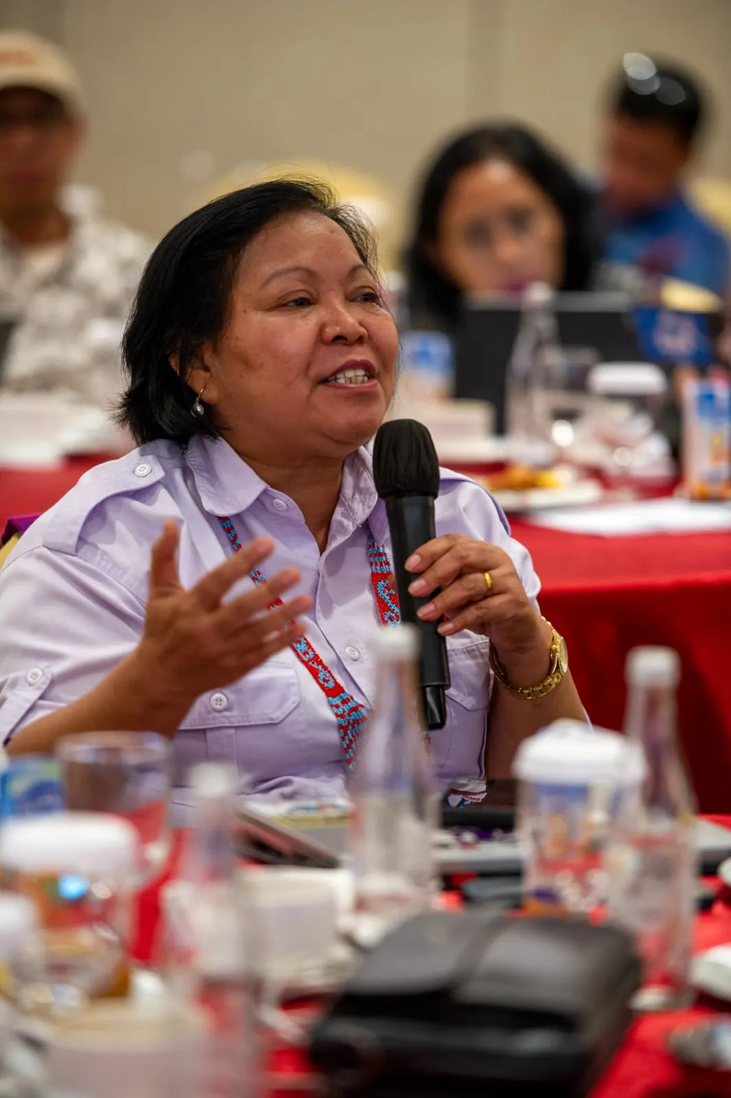 A woman in a lilac shirt with beaded trim speaking into a microphone from the floor of the room
