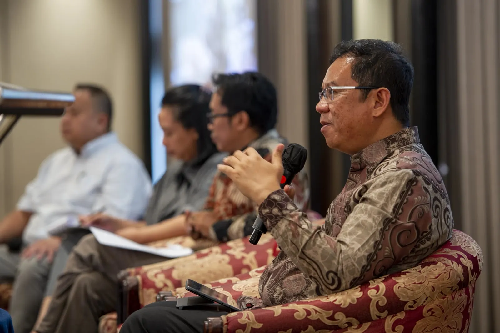A panellist in patterned batik speaking into a microphone, other panellists out of focus beside him