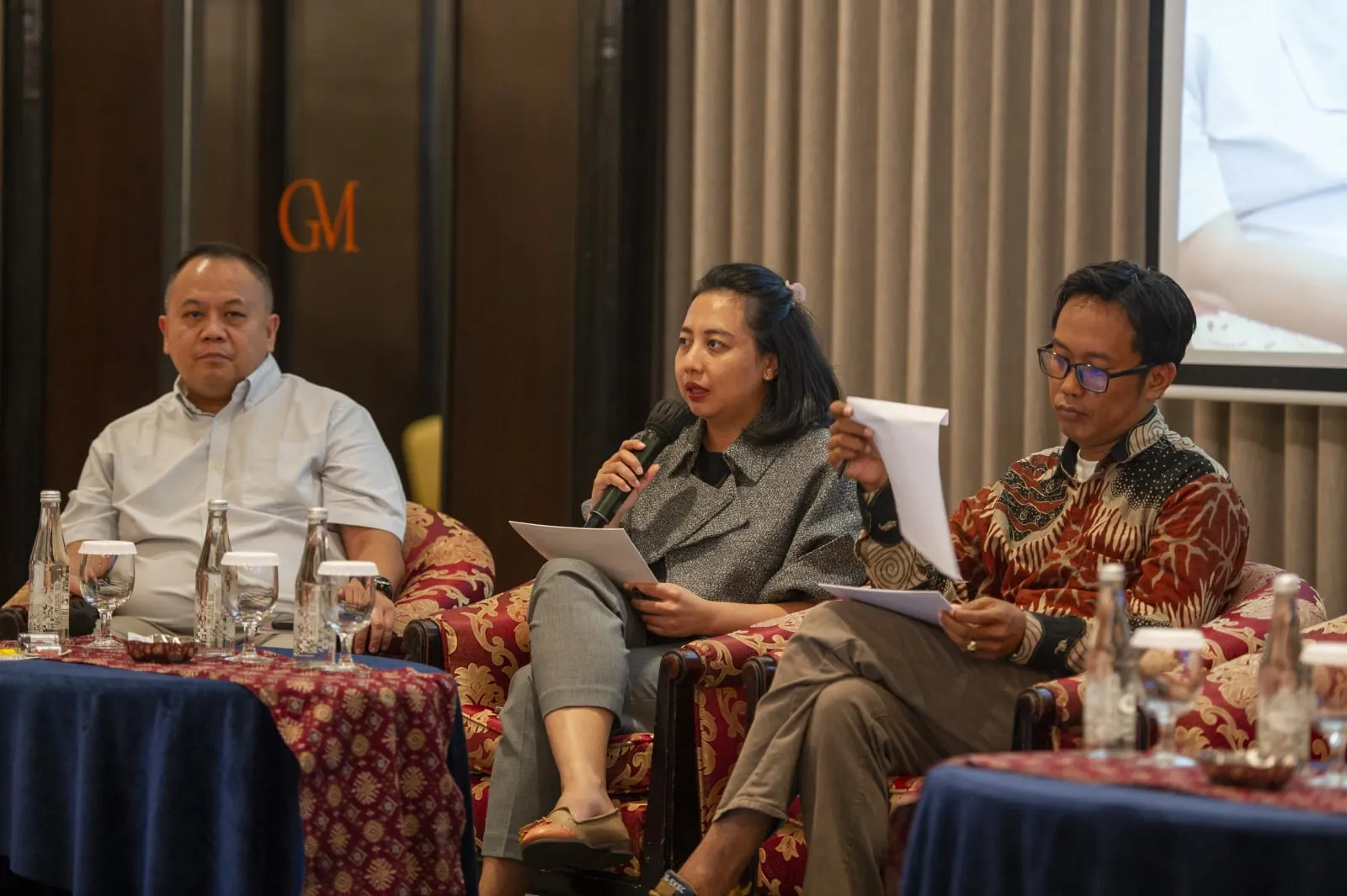 Three panellists seated in armchairs, the woman in the centre speaking into a microphone while holding papers