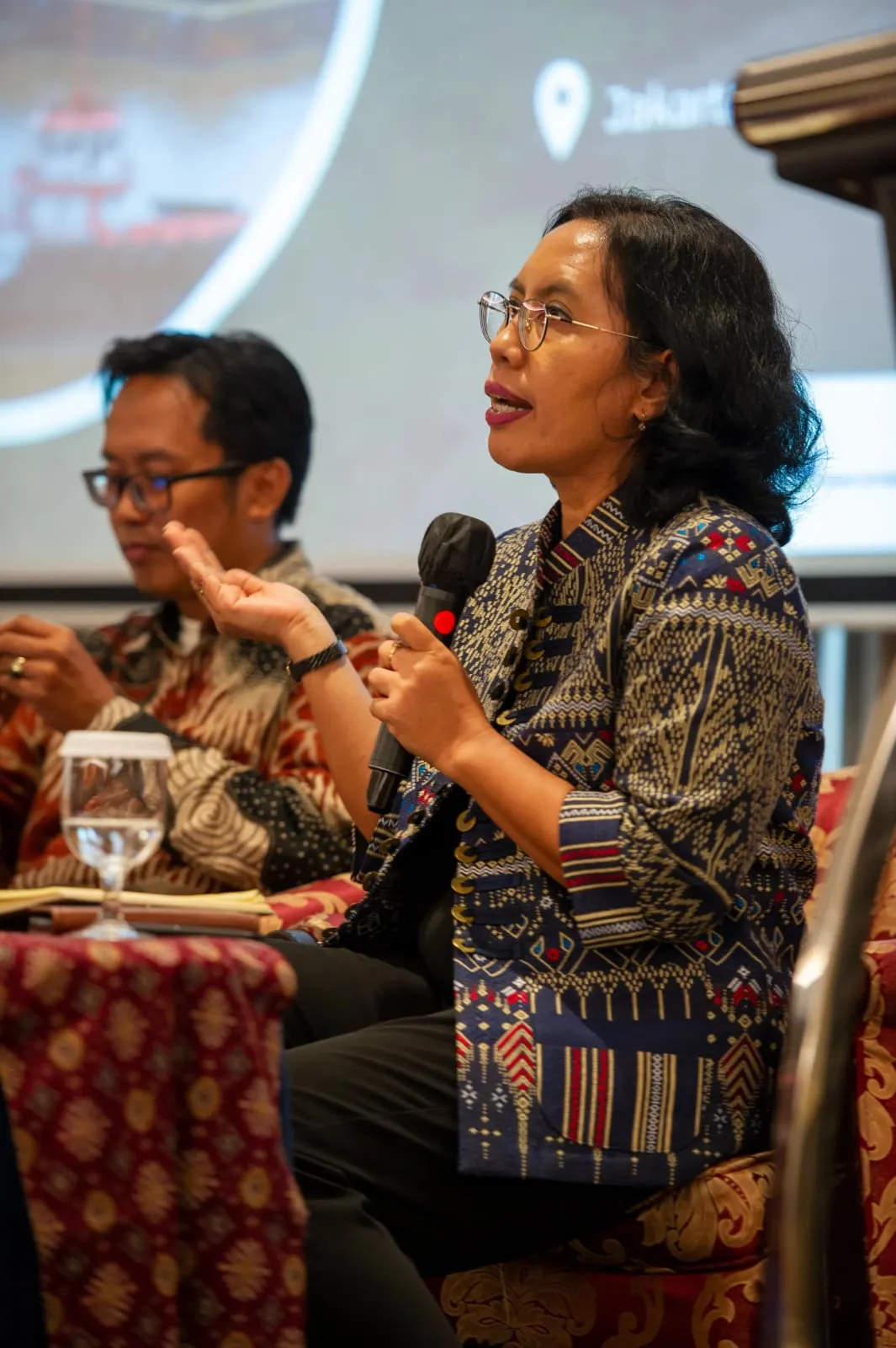 A woman in a blue ikat jacket speaking into a microphone, a colleague listening beside her
