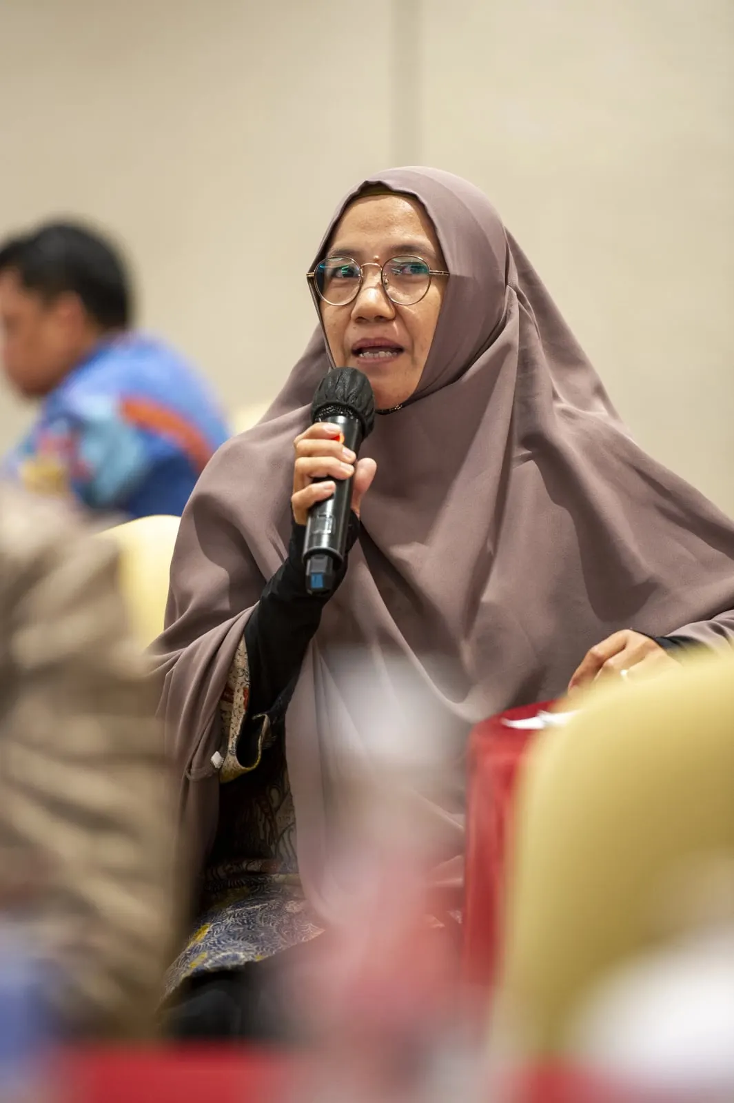 A woman in a mauve headscarf speaking into a microphone from among the seated participants