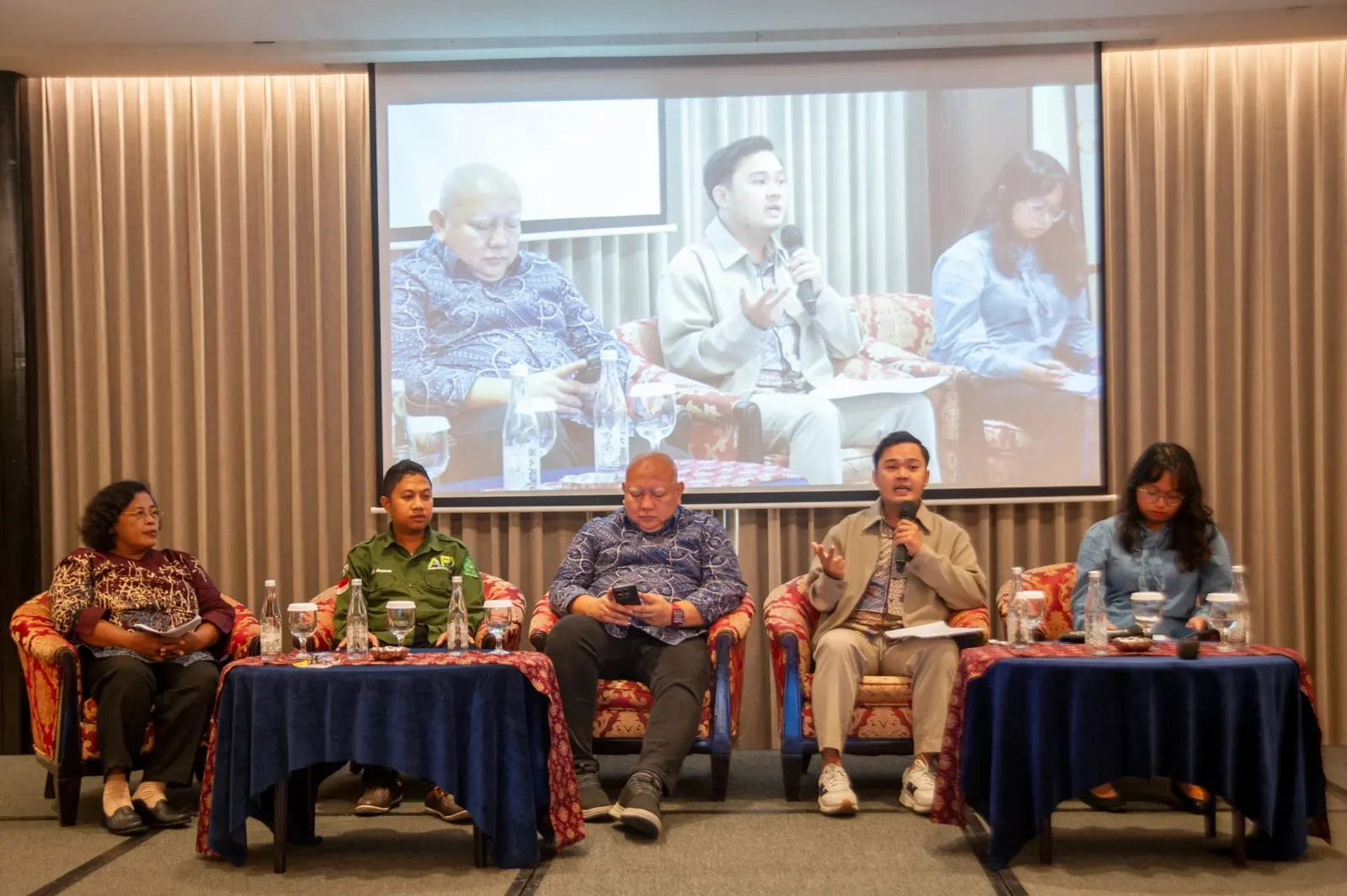 Five panellists on stage, one speaking, the moment relayed on the large screen behind them