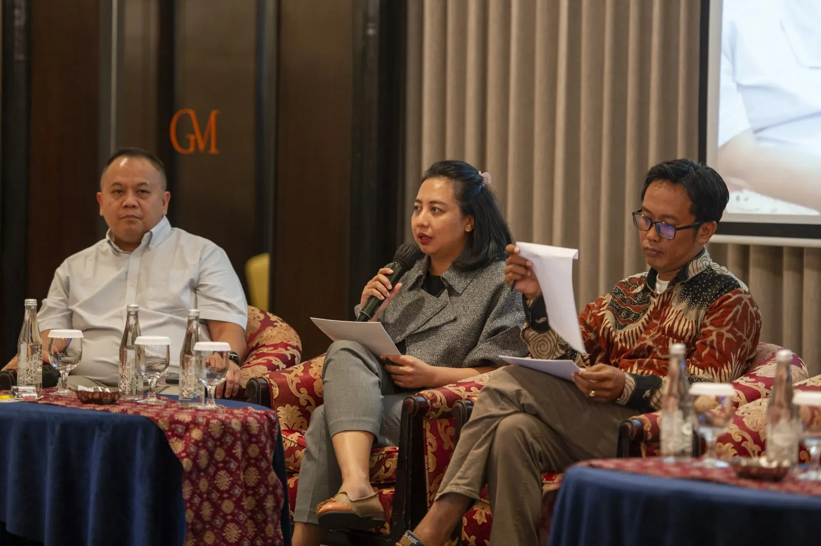 Three panellists seated in armchairs at the national policy dialogue, the woman in the centre speaking into a microphone while holding a sheaf of papers
