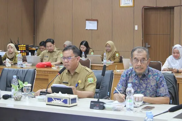 Two men seated at the meeting table, one in government khaki speaking, with participants along the tables behind