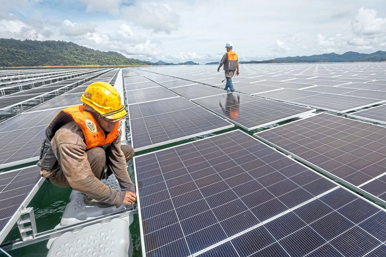 A worker in a yellow hard hat and orange vest crouches on a floating solar farm as a second worker walks across the panels, with forested hills behind