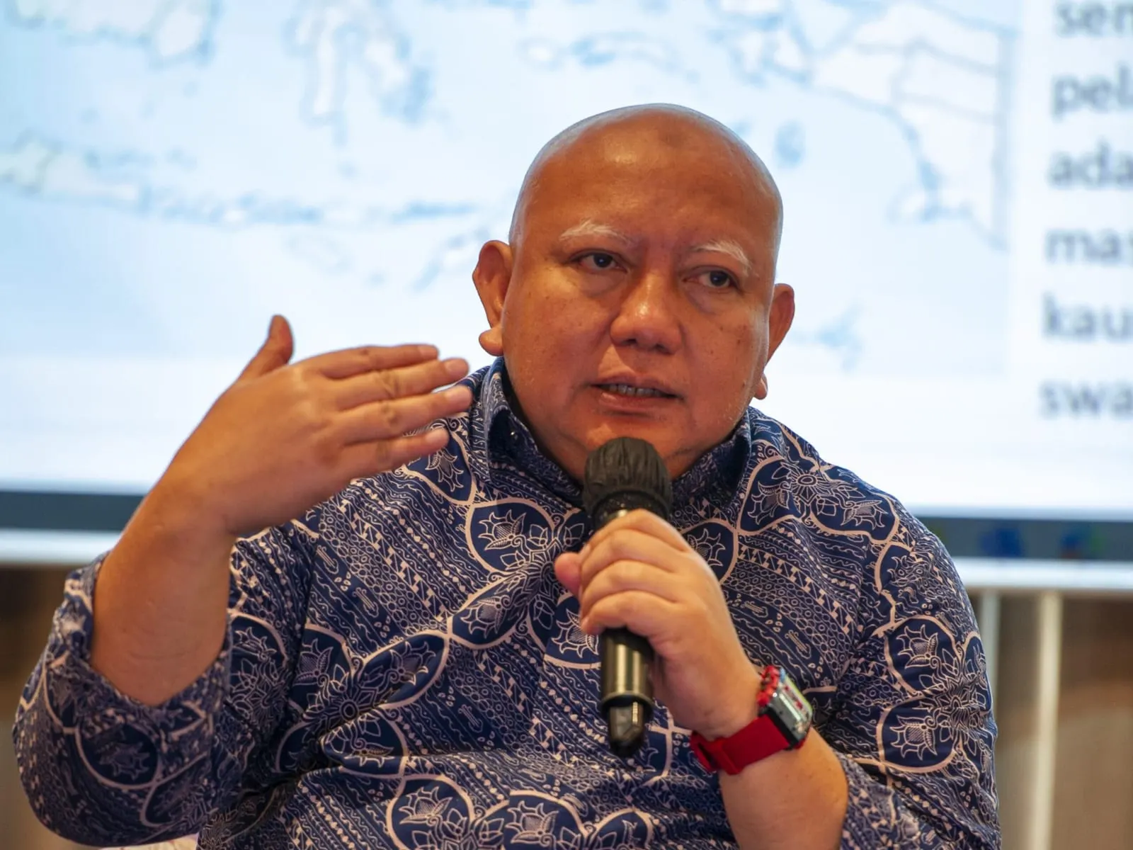Dicky Edwin Hindarto of Yayasan Mitra Hijau speaking into a handheld microphone at a community session, wearing a blue batik shirt