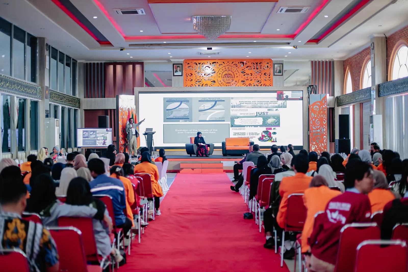 A packed forum hall with delegates seated in rows facing a speaker on stage, a wide screen behind showing Just Transition Forum Asia slides