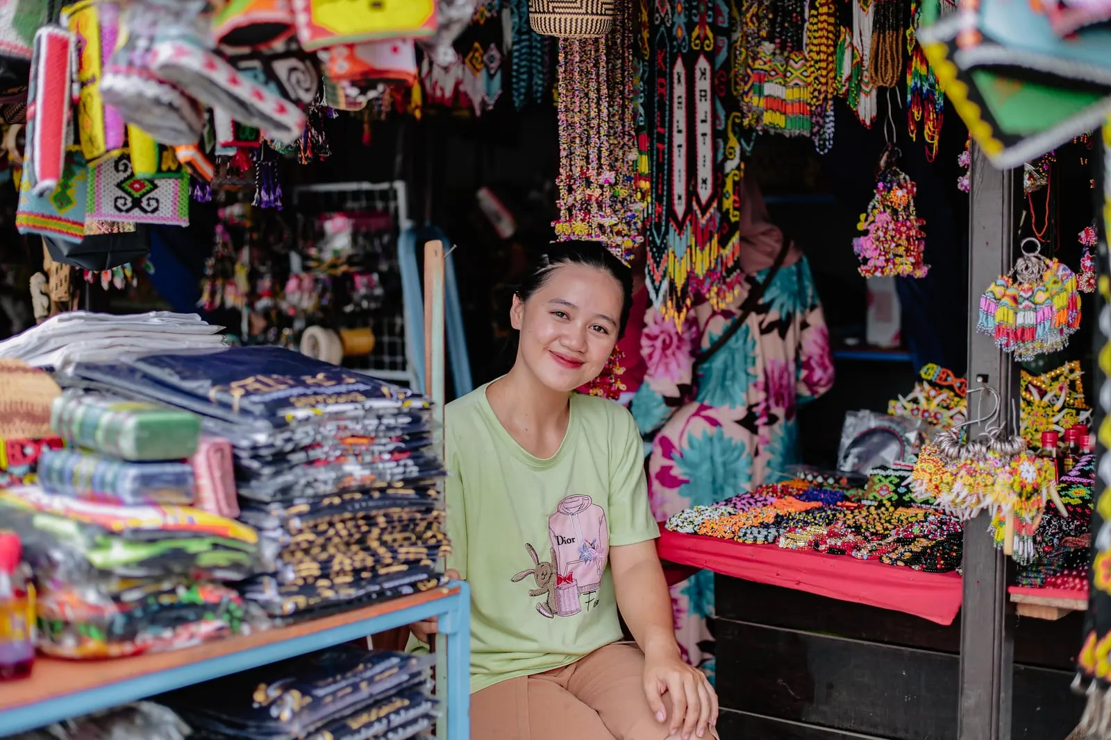 A young woman sitting in a brightly stocked market stall hung with beaded jewellery, embroidered bags and woven crafts