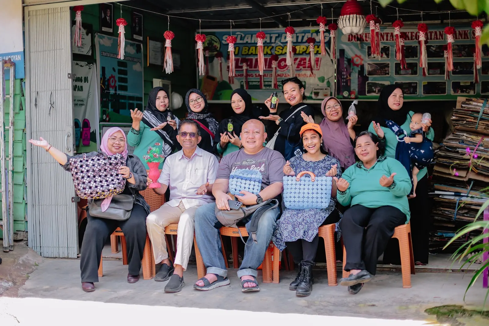 Members of a waste bank collective, mostly women, gathered for a group photograph outside their sorting shed, holding bottled products and bags made from recycled materials