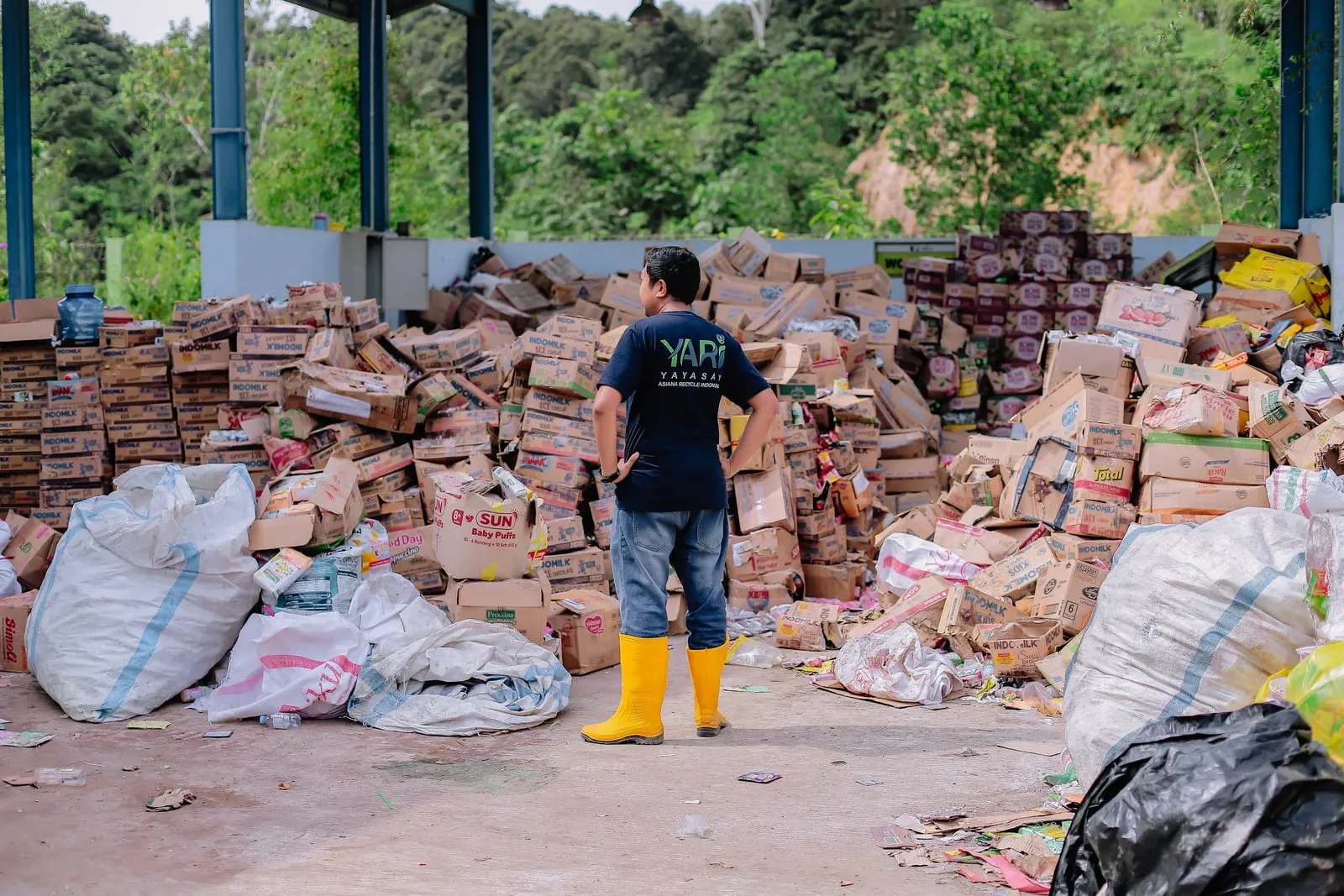 A worker in a recycling foundation T-shirt and yellow boots standing before a large sorted pile of cardboard and packaging at a waste facility