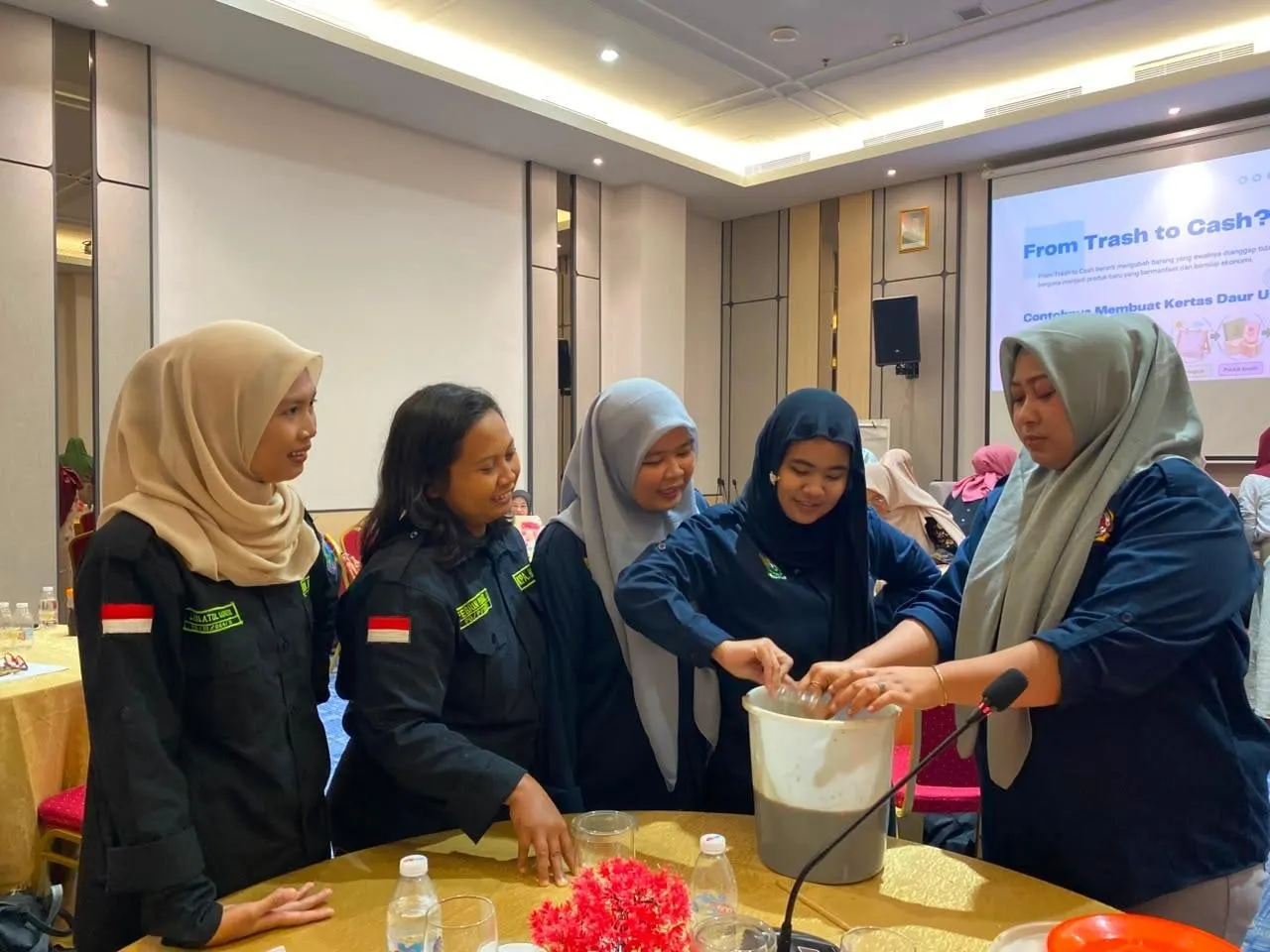 Five women in dark field jackets gathered around a bucket at a training table, mixing eco-enzyme, a From Trash to Cash slide projected behind them