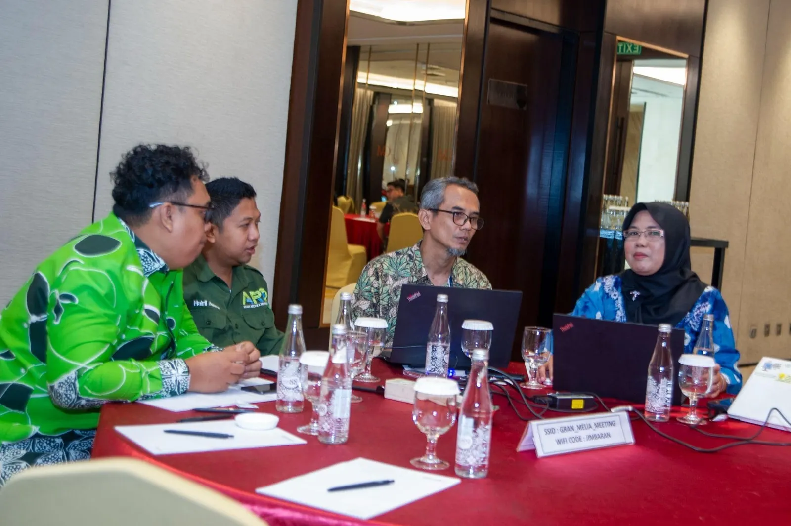 Ari Firmansyah in a green batik jacket in conversation at a round table during the national policy dialogue, delegates and laptops along the table