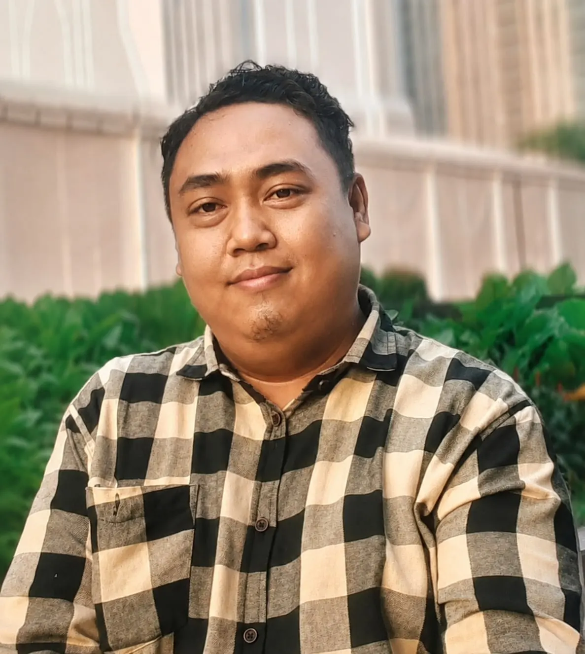Ari Firmansyah outdoors in a black and cream checked shirt, greenery behind him