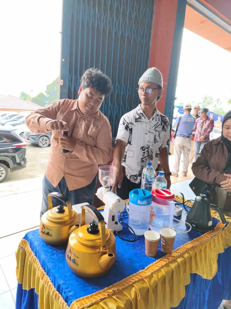 Ari grinding coffee at a stall set with brass kettles and brewing equipment, a colleague beside him