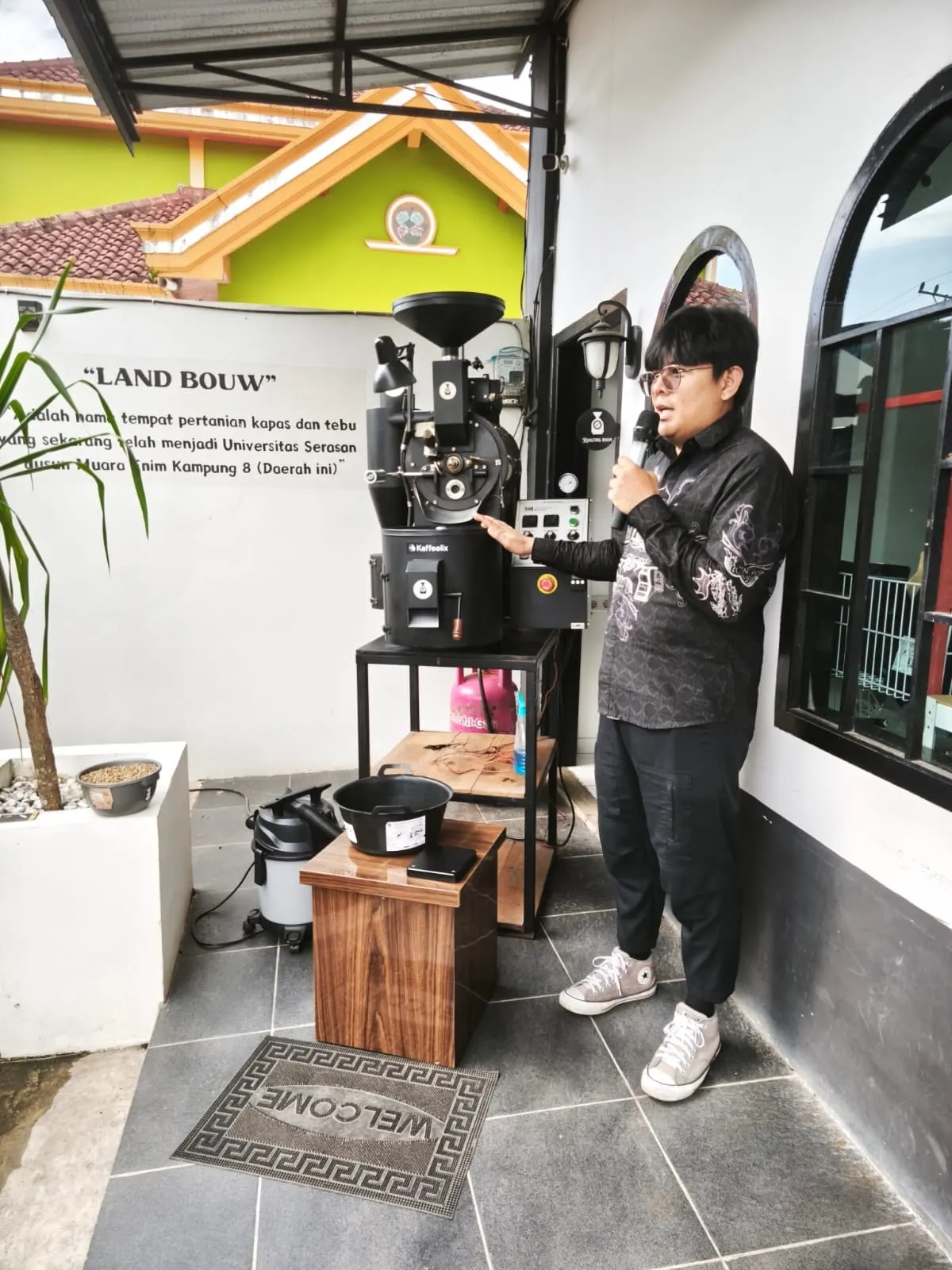 Robby standing beside a drum coffee roaster, speaking into a microphone and gesturing towards the machine