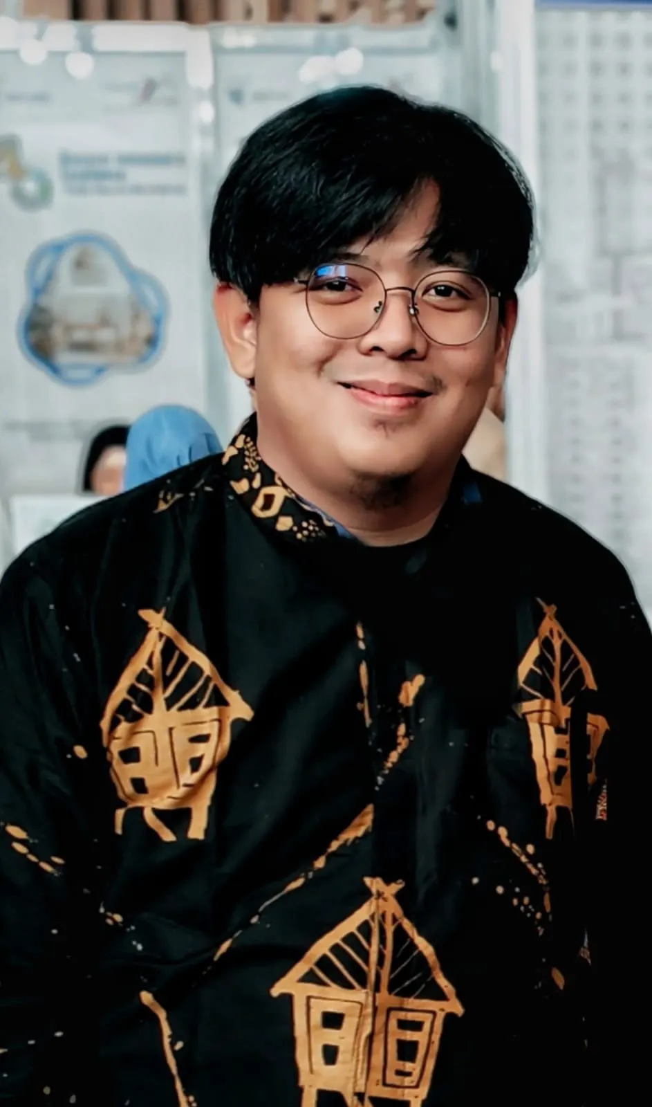 Robby Anugrah Putra, wearing glasses and a black batik shirt patterned with traditional stilt houses, smiling at the camera