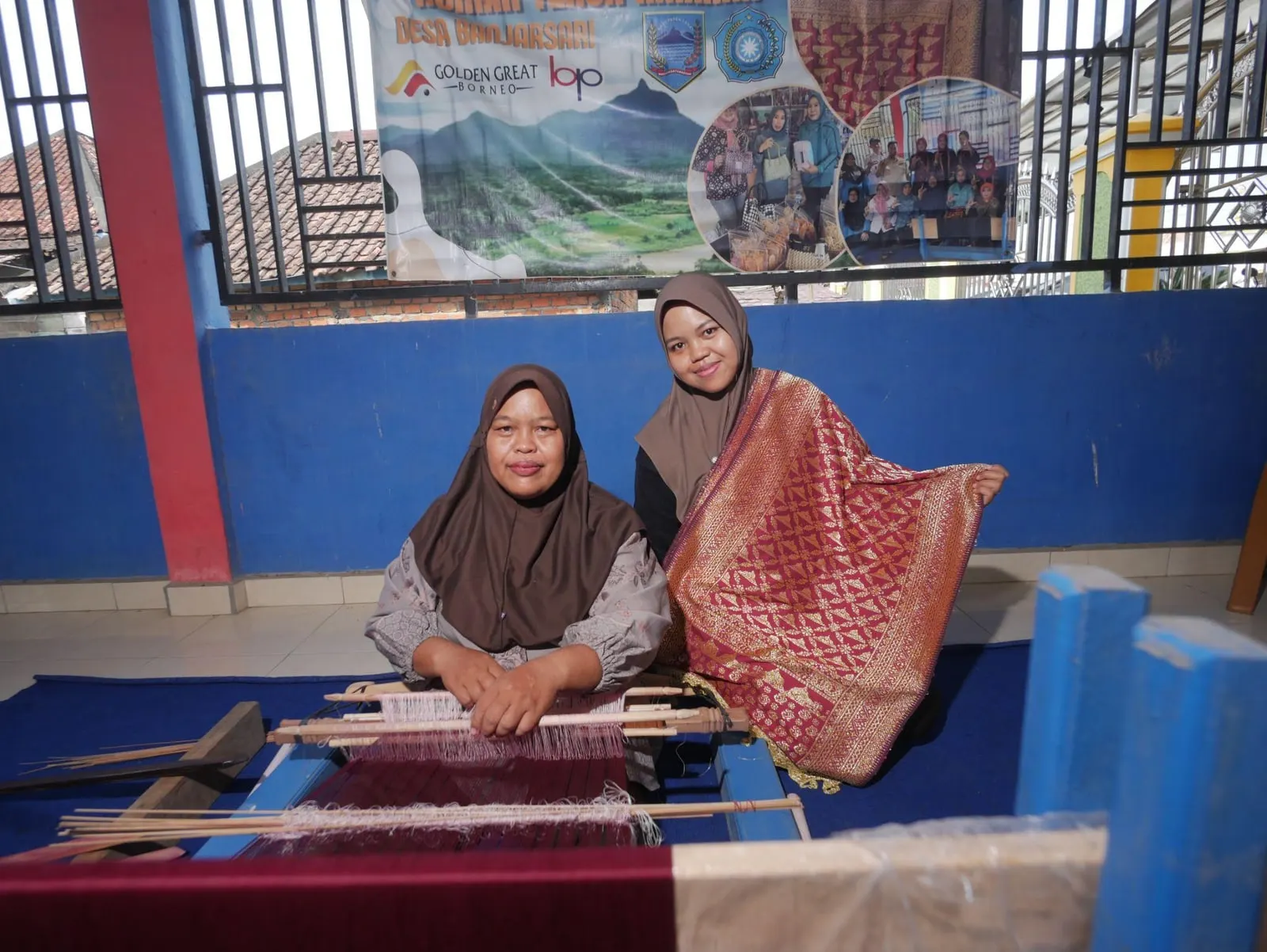 Ibu Rozita seated at her handloom beside her daughter, who holds up a finished crimson and gold songket