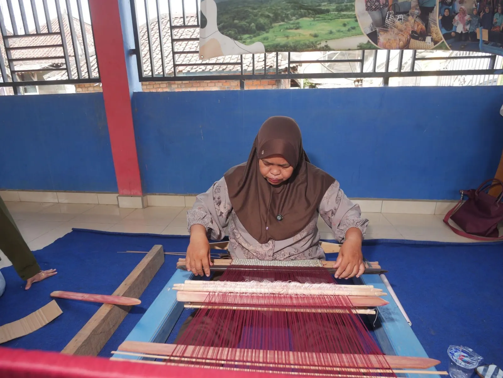 Ibu Rozita working at a low wooden handloom, crimson silk threads stretched before her