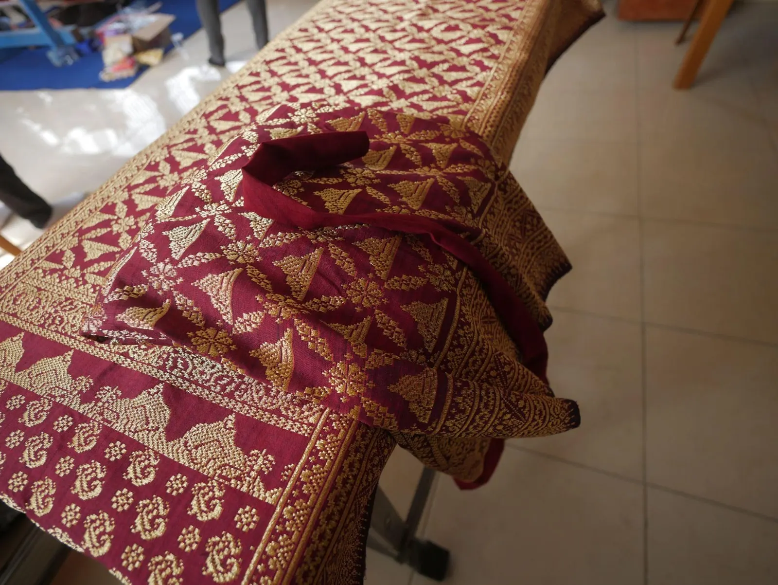 A finished songket folded over a rail, its crimson silk worked in dense gold thread with repeating peaked motifs