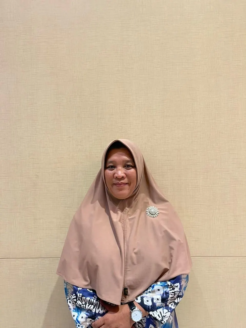 Ibu Ani Tasriah standing against a plain wall, in a pale headscarf and patterned blue dress