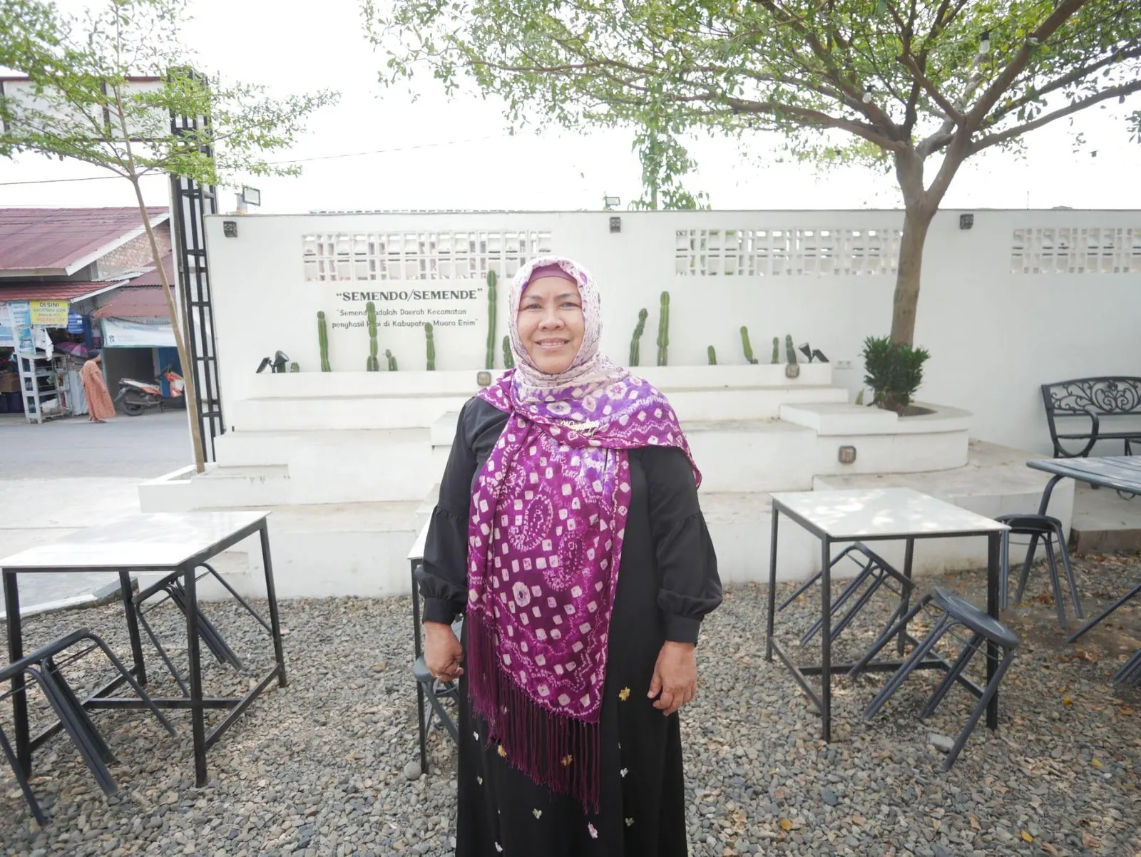 Ibu Nurmalina standing in a courtyard before a white wall lettered “SEMENDO/SEMENDE”, wearing a purple patterned shawl